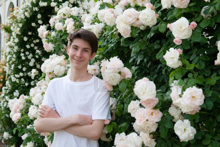 Foto de Adolescente chico en parque entre hermosas rosas florecientes de color blanco-rosa, verano al aire libre - Imagen libre de derechos