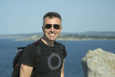 Téléchargez les photos : Homme d'âge moyen avec lunettes de soleil et sac à dos heureux souriant à la côte, portrait d'été en plein air. Un bel homme mûr. Portrait masculin extérieur. - en image libre de droit