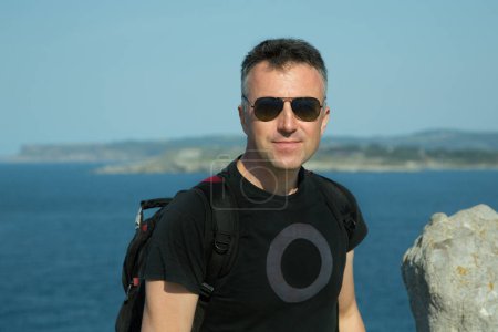 Téléchargez les photos : Homme d'âge moyen avec lunettes de soleil et sac à dos heureux souriant à la côte, portrait d'été en plein air. Un bel homme mûr. Portrait masculin extérieur. - en image libre de droit