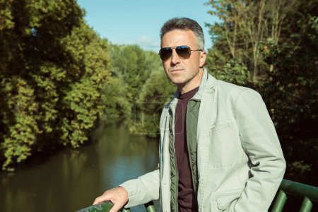 Téléchargez les photos : Homme d'âge moyen avec des lunettes de soleil posant au bord de la rivière, portrait d'automne en plein air. Un bel homme mûr. Portrait masculin extérieur. - en image libre de droit