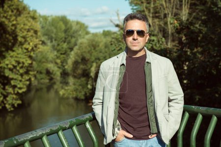 Téléchargez les photos : Homme d'âge moyen avec des lunettes de soleil posant au bord de la rivière, portrait d'automne en plein air. Un bel homme mûr. Portrait masculin extérieur. - en image libre de droit