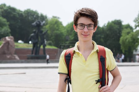 Téléchargez les photos : Jeune beau garçon adolescent de 15 ans portant un t-shirt jaune avec un sac à dos regardant la caméra et souriant heureux, parc d'été en plein air - en image libre de droit