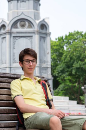 Téléchargez les photos : Jeune beau garçon adolescent de 15 ans portant un t-shirt jaune assis sur un banc dans un parc d'été - en image libre de droit