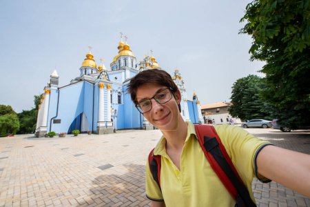 Photo for Young handsome 15 years old teen boy looking at camera and happy smiling takes a selfie with smartphone over Mikhailovsky monastery, summer city centr of Kyiv, Ukraine - Royalty Free Image