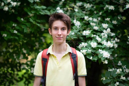 Photo for Young handsome 15 years old teen boy wearing yellow t-shirt with backpack looking at camera and happy smiling, summer park outdoor with blooming flowers - Royalty Free Image
