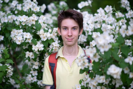 Téléchargez les photos : Jeune beau garçon adolescent de 15 ans portant un t-shirt jaune avec un sac à dos regardant la caméra et souriant heureux, parc d'été en plein air avec des fleurs en fleurs - en image libre de droit