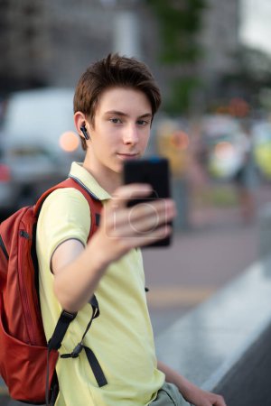 Foto de Joven chico guapo con auriculares toma selfie con teléfono inteligente, parque de verano al aire libre. Adolescente de 15 años usando teléfono móvil, estilo de vida juvenil - Imagen libre de derechos