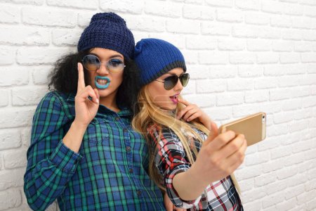 Foto de Dos chicas divertidas felices tomando selfie con teléfonos inteligentes sobre la pared de ladrillos blancos - Imagen libre de derechos