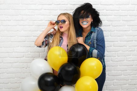 Téléchargez les photos : Deux filles hipster heureux dans des lunettes de soleil s'amusent avec des ballons sur fond de mur blanc. - en image libre de droit