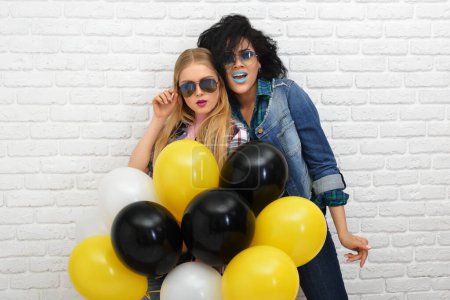 Photo for Two happy hipster girls in sunglasses have fun with balloons over white wall background. - Royalty Free Image