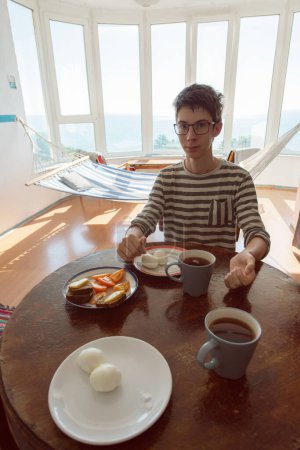 Téléchargez les photos : Beau garçon adolescent mangeant dans une chambre vintage avec hamac et vue sur la mer. Intérieur du XXe siècle, nostalgie - en image libre de droit