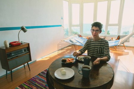 Photo for Handsome teen boy eating in vintage room with old fashioned armchair, retro radio turntable, table with food, telephone, standart lamp, doormat, chairs, hammock and sea view. Interior of 20th century, nostalgia - Royalty Free Image