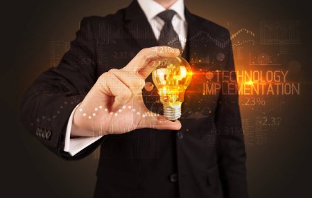 Téléchargez les photos : Homme d'affaires tenant une ampoule avec inscription TECHNOLOGY IMPLEMENTATION, concept de technologie d'entreprise - en image libre de droit