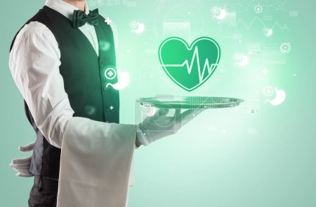 Photo for Handsome young waiter in tuxedo holding tray with ECG icons on tray, global healthcare concept - Royalty Free Image