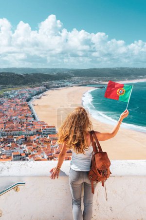 Foto de Mujer con bandera portuguesa- Famosa playa de Nazare- estremadura, Leiria - Portugal- vacaciones, destino turístico, concepto de turismo - Imagen libre de derechos