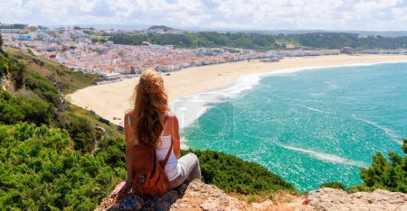 Téléchargez les photos : Femme touriste assis sur la falaise et bénéficiant d'une vue panoramique sur l'océan Atlantique, plage Nazare au Portugal - en image libre de droit