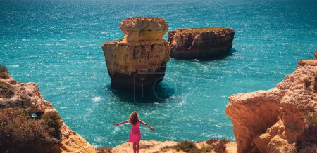 Foto de Mujer joven con vestido rojo disfrutando de vista panorámica de la costa atlántica del Algarve, Turismo en Portugal- destino de viaje de verano, mujer activa saludable, concepto de bienestar - Imagen libre de derechos