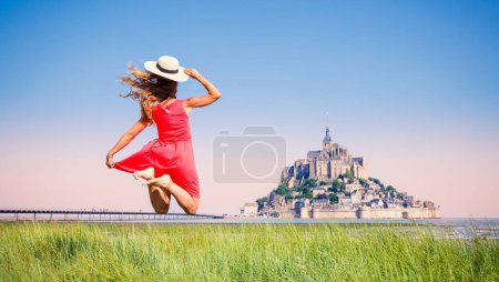 Foto de Mujer feliz con vestido rojo y sombrero saltando frente al Mont Saint Michel- Normandie en Francia - Imagen libre de derechos