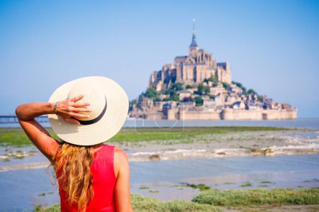 Foto de Vista trasera de la mujer con vestido rojo y sombrero mirando Le Mont Saint Michel- Normandía en Francia - Imagen libre de derechos
