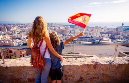 Frau und Kinder mit spanischer Nationalflagge blicken auf andalusische Stadtsilhouette