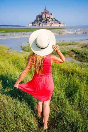 Foto de Atractiva mujer con vestido rojo y sombrero disfrutando de la vista del Mont Saint Michel- Normandía en Francia- destino de viaje, turismo turístico, concepto de fantasía - Imagen libre de derechos