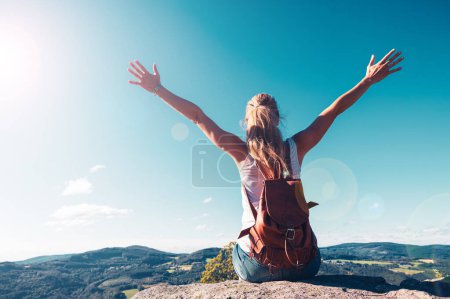 Woman sitting on a peak with backpack and arms raised against blue sky- freedom, achievement,travel,adventure concept