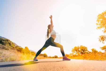 Téléchargez les photos : Jeune femme faisant du yoga dans la nature sur la route du lever du soleil sport, flexibilité, mode de vie actif, concept d'étirement - en image libre de droit