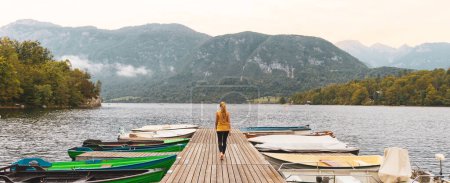 Téléchargez les photos : Femme touriste marchant sur la jetée avec des bateaux sur le lac Bohinj en Slovénie- Voyage, vacances, tourisme d'excursion - en image libre de droit