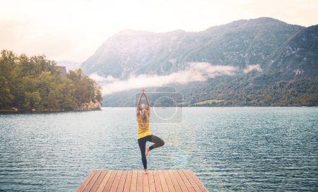Téléchargez les photos : Femme faisant des exercices de yoga sur jetée, lac et montagne le matin- sport, relaxation, méditation - en image libre de droit