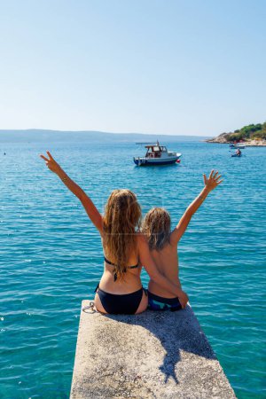 Téléchargez les photos : Mère et fils assis sur la jetée profitant de la mer et le bateau Voyage, vacances, concept de vacances en famille - en image libre de droit