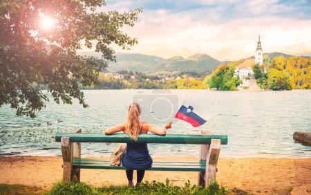 Foto de Mujer sentada en el banco con bandera eslovena disfrutar de la vista de la iglesia de peregrinación en el lago Bled-viajes, turismo turístico, vacaciones en Eslovenia - Imagen libre de derechos