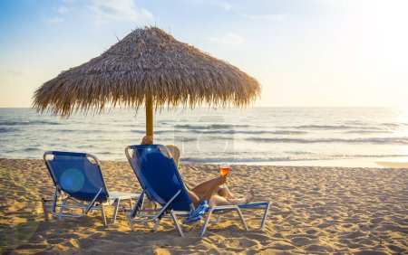 Foto de Playa resort, chaise lounge y sombrilla en la arena frente al mar- Mujer relajándose con una copa de vino al atardecer - Imagen libre de derechos