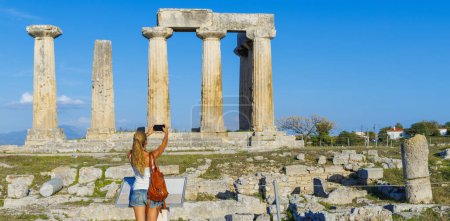 Foto de Templo de Apolo, ruinas del antiguo templo en Corinto - turista mujer tomando fotos con smarpthone- viajes en Grecia, Europa - Imagen libre de derechos