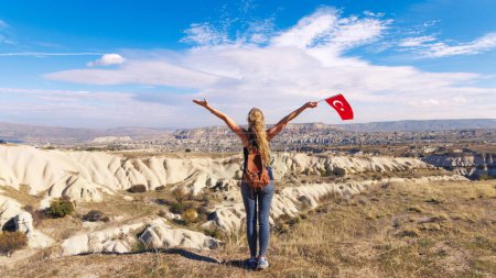 Foto de Mujer mochilera feliz sosteniendo bandera turca disfrutando de vista panorámica de Capadocia - Imagen libre de derechos