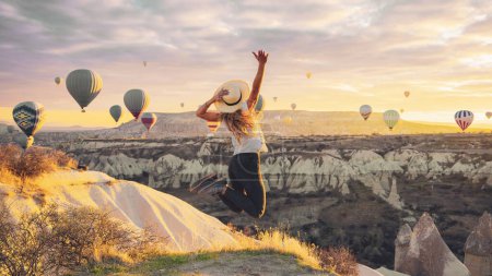 Photo for Young female tourist jumping in the air with hot air balloons background at sunrise in Cappadocia, Turkey - Royalty Free Image