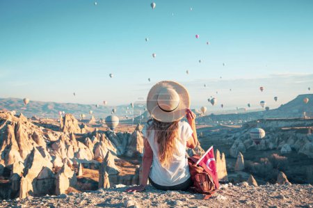 Foto de Kapadoya Capadocia en Turquía- turista joven hembra sentada y viendo coloridos globos de aire caliente sobre el famoso paisaje de chimeneas en Capadocia al amanecer- Destino de viaje, vacaciones, turismo turístico en Turquía - Imagen libre de derechos