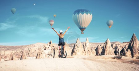 Téléchargez les photos : Femme touriste en vélo profitant du paysage de la Cappadoce avec des montgolfières - en image libre de droit