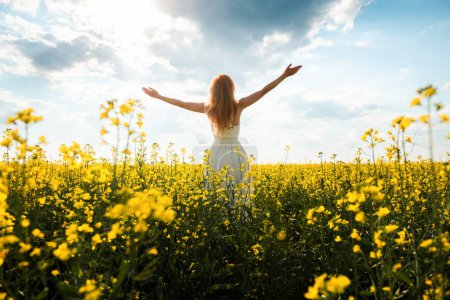 Téléchargez les photos : Femme en robe longue se tient dans un champ avec des fleurs jaunes et levant les mains au soleil. Paysage estival. Concept de plaisir et de bonheur - en image libre de droit