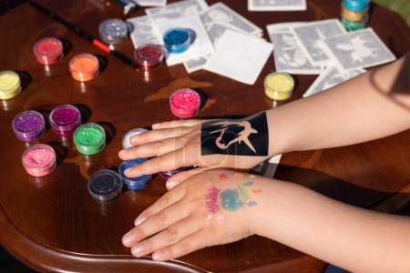 Foto de El animador del niño está creando un brillante tatuaje de purpurina en la mano de un niño en una fiesta de cumpleaños. Arte corporal - Imagen libre de derechos