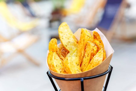 Téléchargez les photos : Oven baked potato wedges with sea salt and herbs. fries in a recyclable paper bag on a blurred street background, popular fast street food - en image libre de droit
