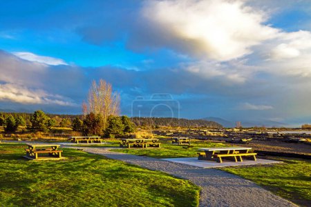 Téléchargez les photos : Au début du printemps, heure du coucher du soleil, aire de pique-nique près du rivage, tables de pique-nique sur une pelouse verte contre une chaîne de montagnes et ciel nuageux orageux. Ce parc est situé sur l'île Sea, près de l'aéroport de Vancouver. - en image libre de droit