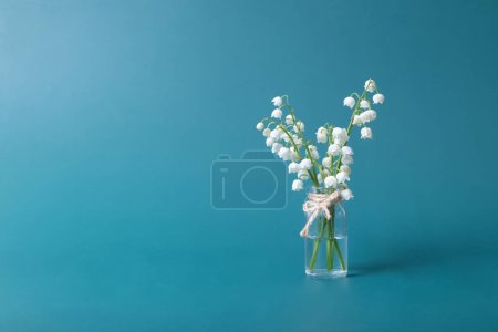 Téléchargez les photos : Lilies of the valley bouquet in a mini glass jar on a turquoise background with copy space. Minimalistic spring still life. - en image libre de droit