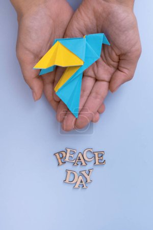 Peace day text from wooden letters with yellow-blue paper dove in hands on blue background top view. International Peace day concept.