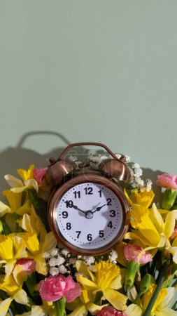 Foto de Reloj despertador con flores primaverales en vista superior de fondo de color, disposición plana. Primavera, concepto de ahorro de luz natural, primavera hacia adelante. - Imagen libre de derechos