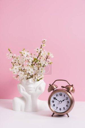 Foto de Reloj despertador y jarrón en forma de cabeza con flores de primavera sobre fondo rosa. - Imagen libre de derechos