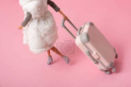 Die Puppe trägt ein Gepäck auf Rädern auf rosa Hintergrund. Reisen, Urlaub kreatives minimalistisches Konzept.