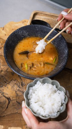 Rice on chopsticks in women's hands on the background of tom yum soup.