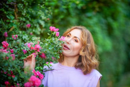 Foto de Hermosa mujer de mediana edad exitosa oliendo rosas florecientes y disfrutando en el jardín al aire libre. Sesión de fotos callejeras - Imagen libre de derechos