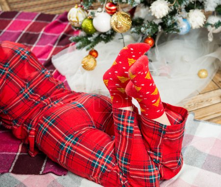 Photo for Young woman in holiday pajamas laying down near christmas tree - Royalty Free Image