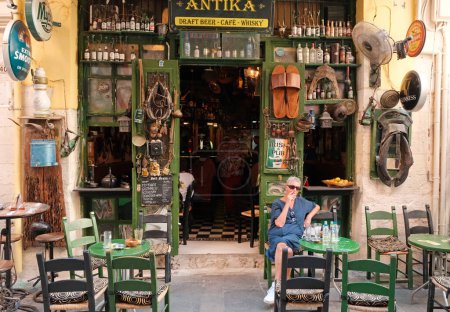 Foto de Rethymnon, Grecia - 09 octubre, 2023: Elegante pub exterior irlandés en la ciudad de Rethymnon en Creta, Grecia con una mujer fumadora solitaria sentada en la mesa al aire libre - Imagen libre de derechos